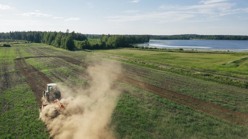 Lantbruks-traktor plogar åkermark med sjö och skog i bakgrunden.